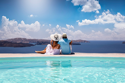 Photo of a couple shot from behind, sitting by a pool looking out to the ocean. 