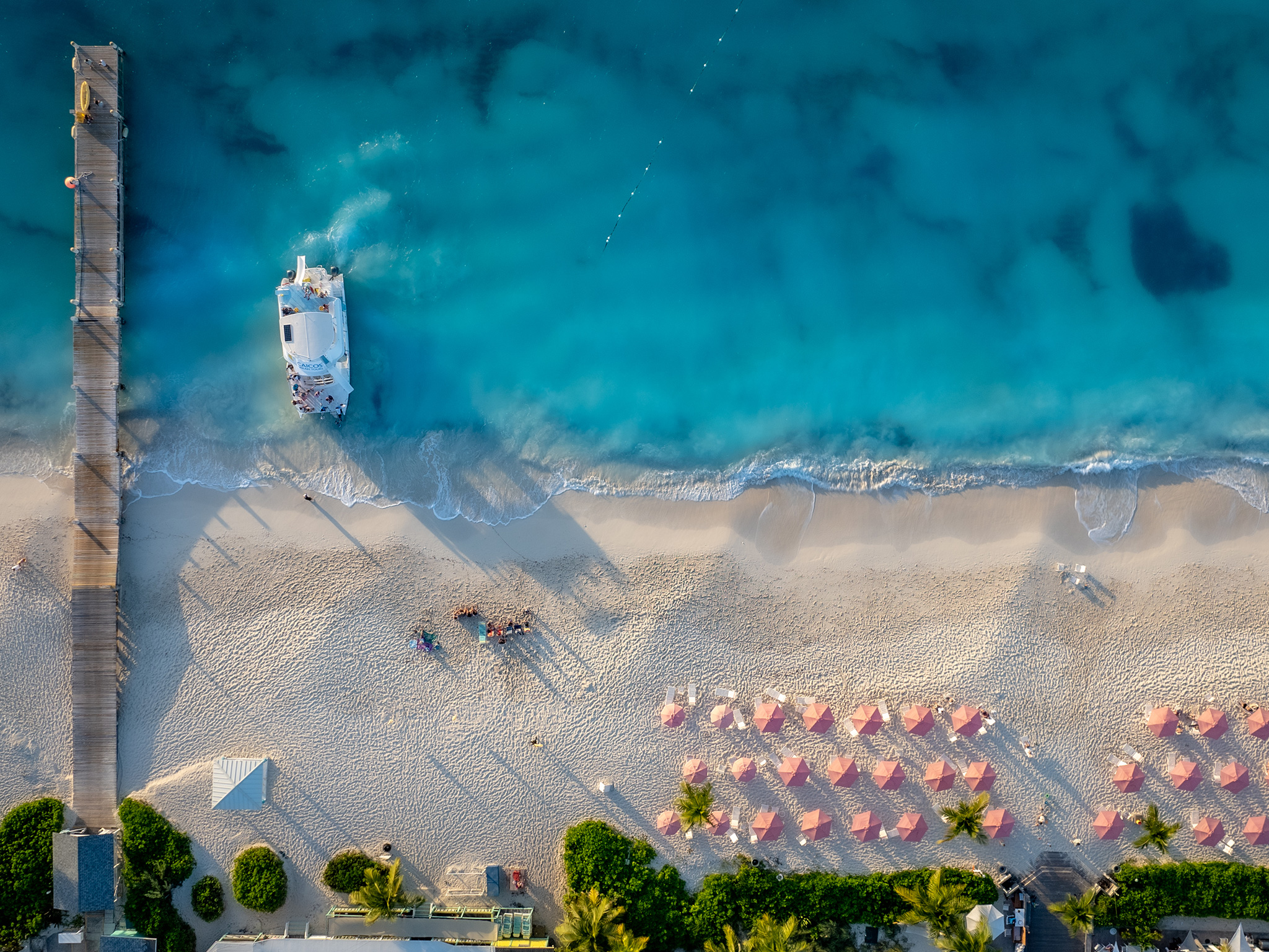 Aerial shot of a beach featuring a boat, pier, and beach umbrellas.